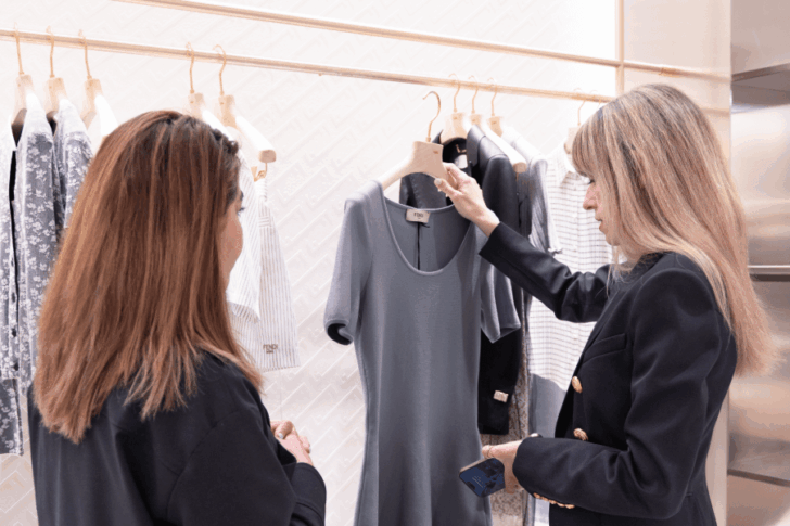 Raquel Macias, personal stylist showing a gray dress to a client inside a modern boutique, with clothing neatly displayed on wooden hangers in the background.
